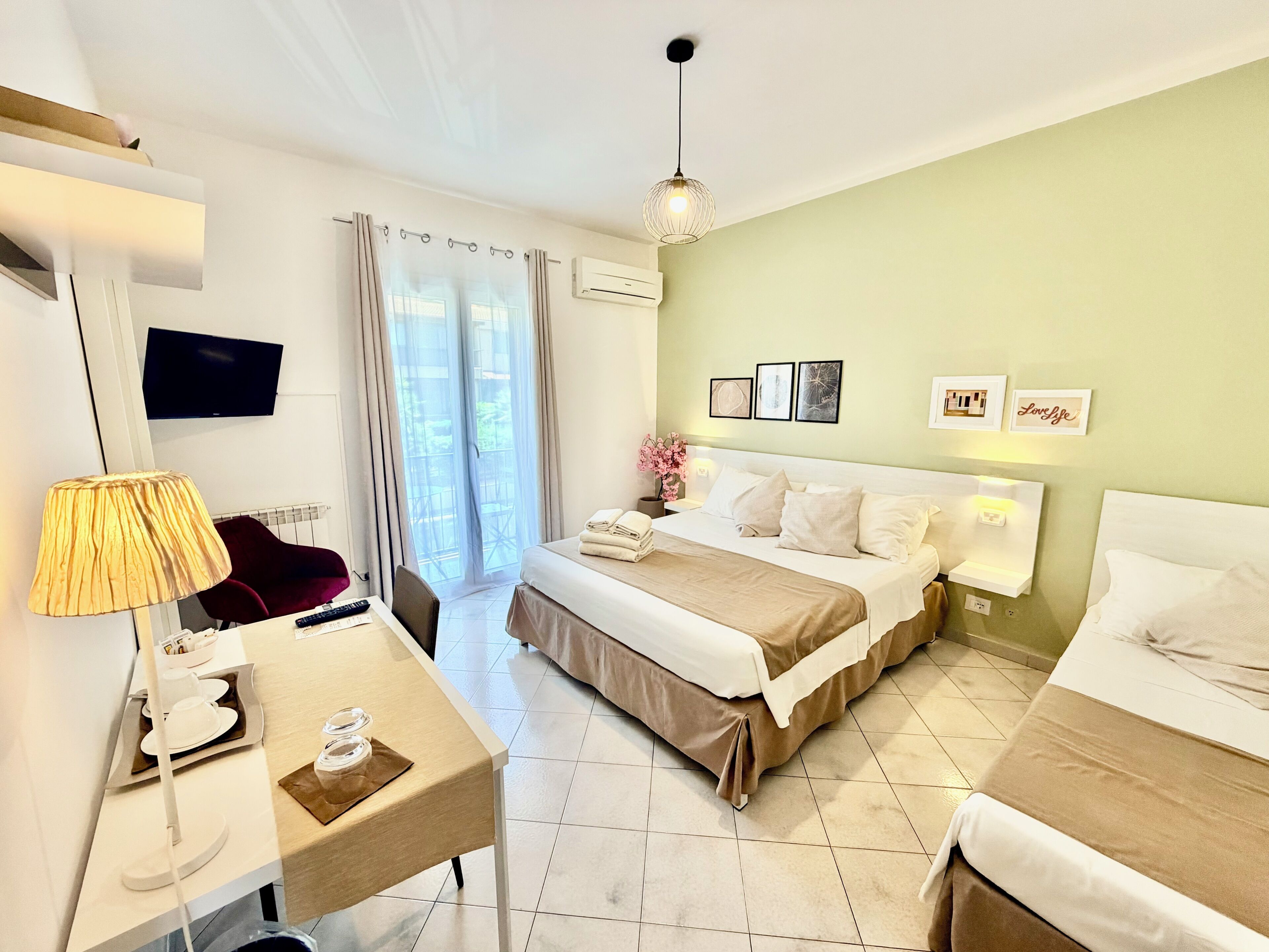 A hotel room with a bed, bedside table, lamp, TV, and a view of the outdoors.