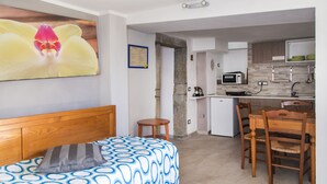 Une chambre à coucher avec un lit, une tête de lit en bois et un oreiller. On trouve un petit tabouret en bois et une table à manger avec des chaises dans la pièce adjacente. La cuisine est équipée d’un four à micro-ondes, d’une bouilloire et d’un évier.