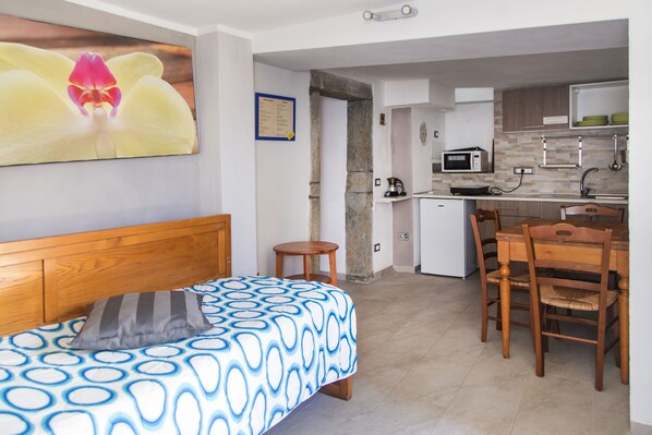 A bedroom with a bed, a wooden headboard, and a pillow. There is a small wooden stool and a dining table with chairs in the adjacent room. The kitchen has a microwave, a kettle, and a sink.