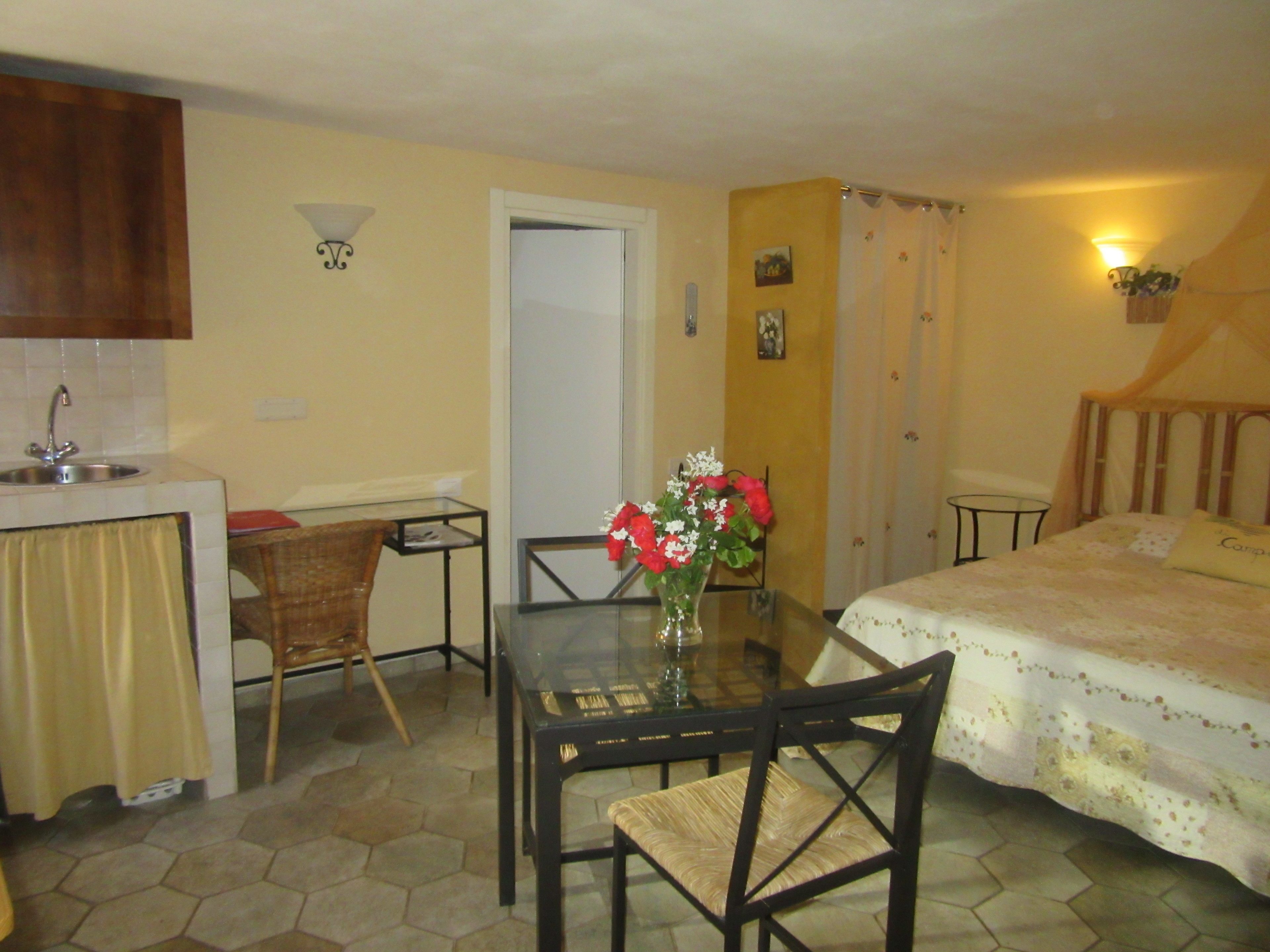 Une pièce de taille modeste comprenant un lit, une table à manger avec des chaises, un petit coin cuisine avec un évier et une porte donnant sur une autre pièce.