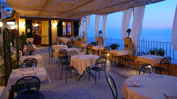 Un espace de restauration en plein air avec des tables et des chaises, une vue sur la mer et une partie abritée avec un toit de chaume.