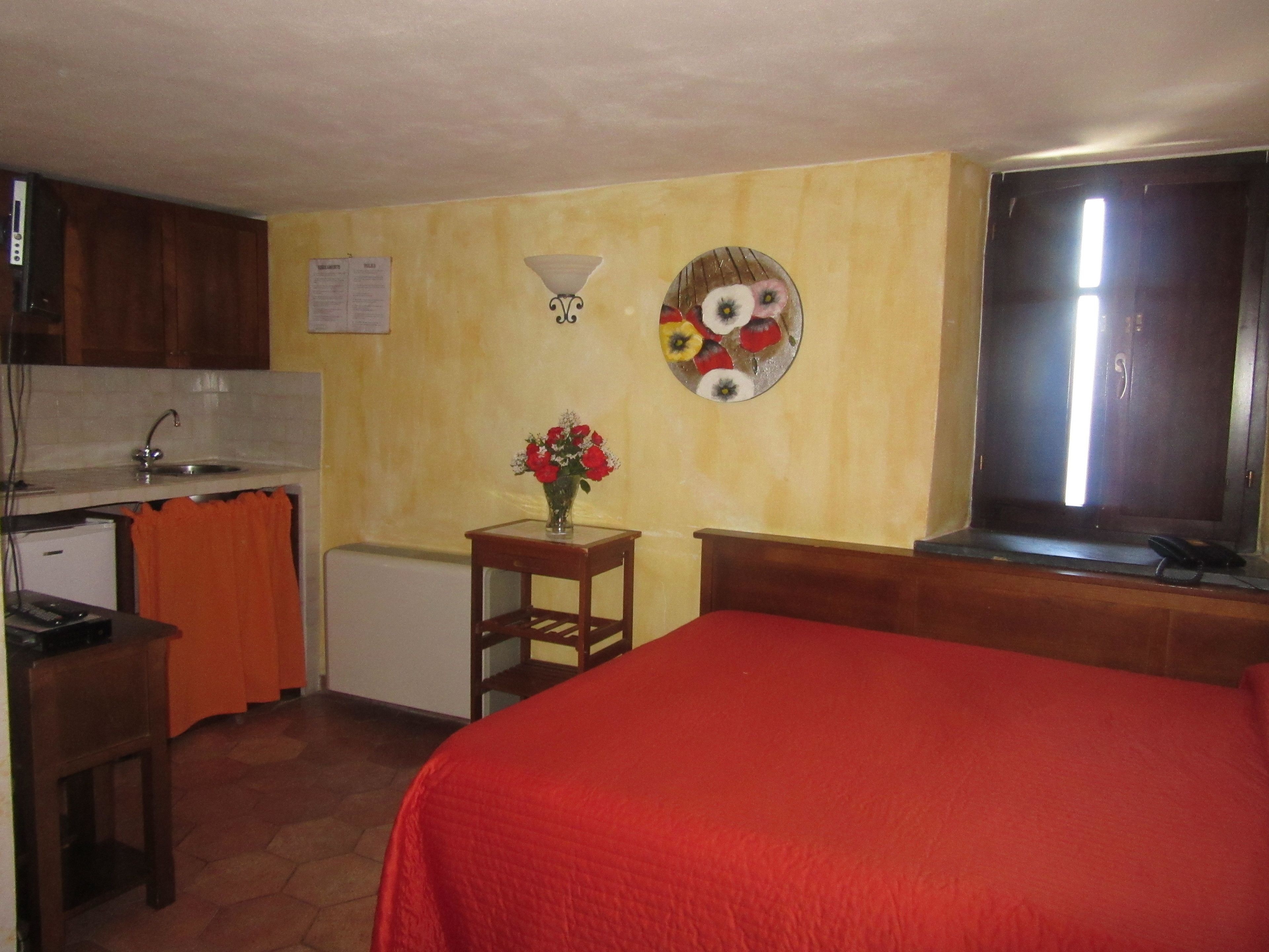 A compact hotel room with a kitchenette, a bed with a red blanket, a small table with a vase of flowers, and a wall-mounted light fixture.