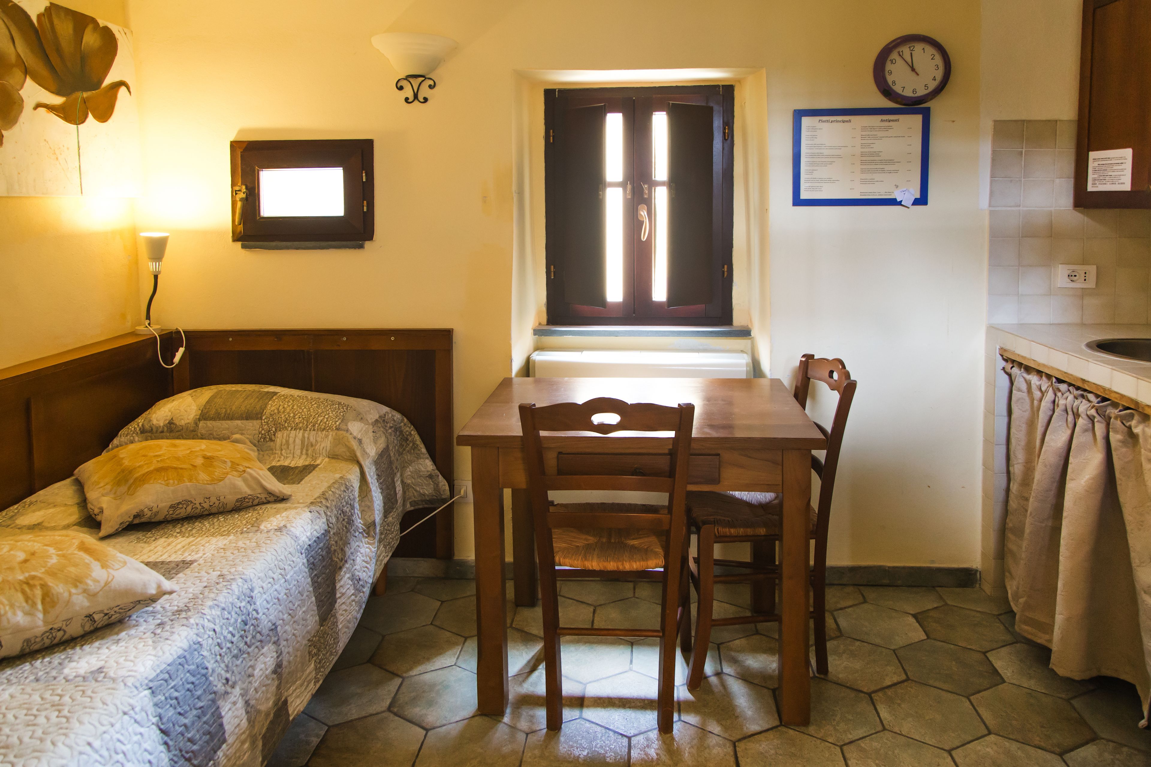 Une petite pièce avec un lit, une table en bois et une chaise. Il y a une fenêtre donnant sur un paysage agréable, une horloge au mur et un tableau d’affichage où est affiché un menu.