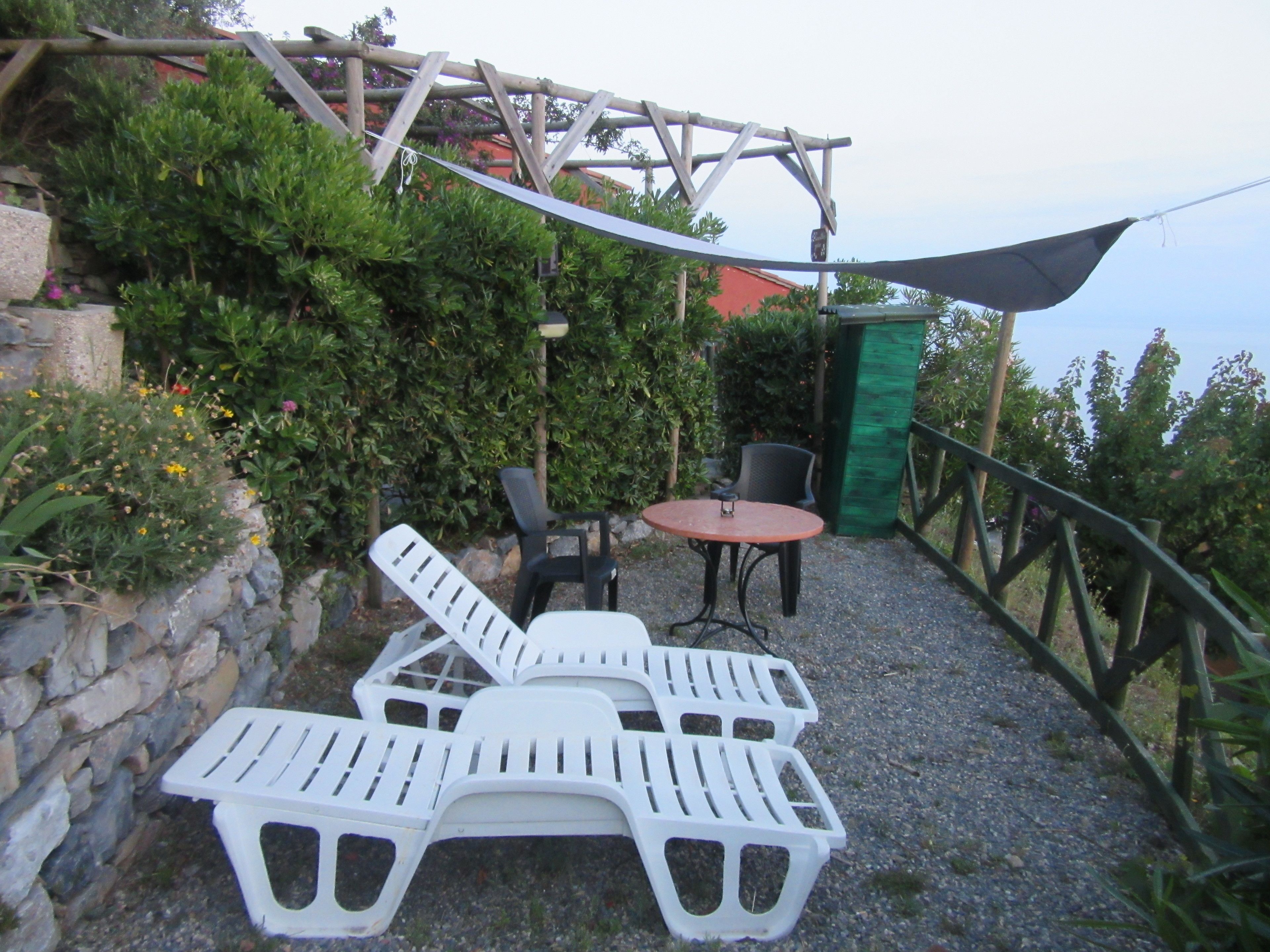 A patio with white plastic lounge chairs and a small round table, surrounded by greenery and a stone wall.