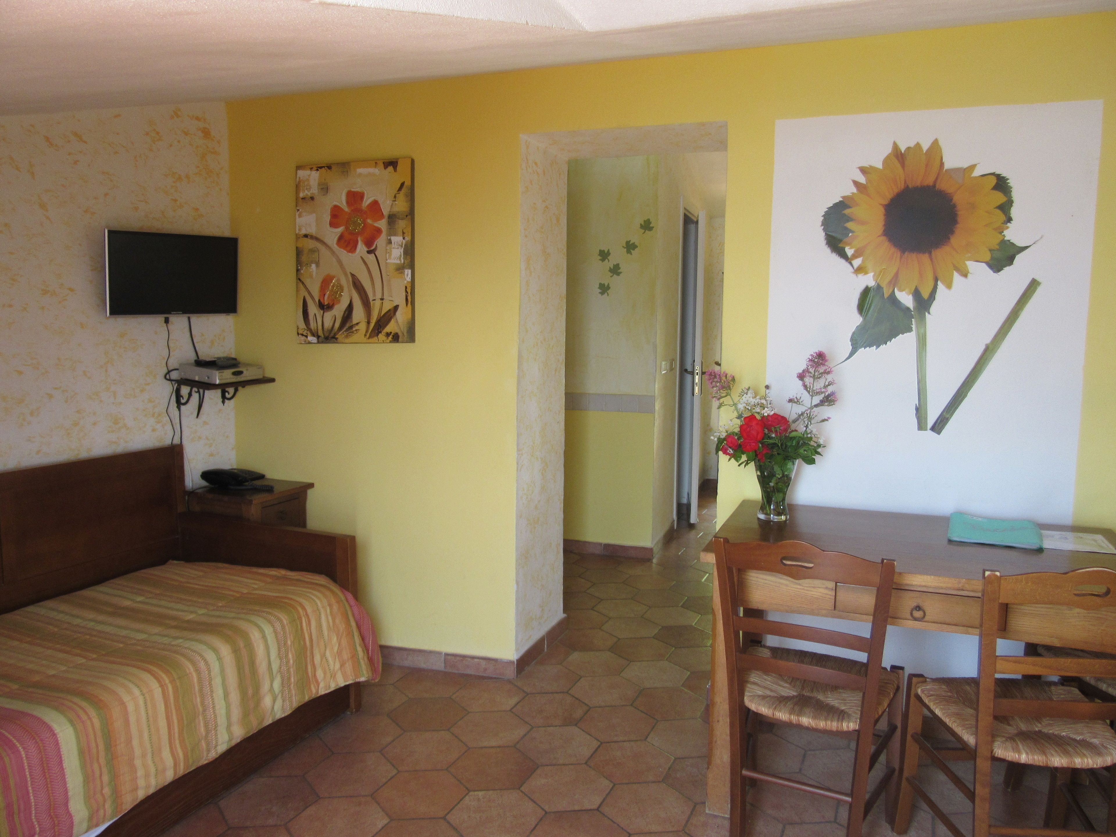 Ein Zimmer mit einem Bett, einem Fernseher, einem Esstisch mit Stühlen und einem Sonnenblumenbild.