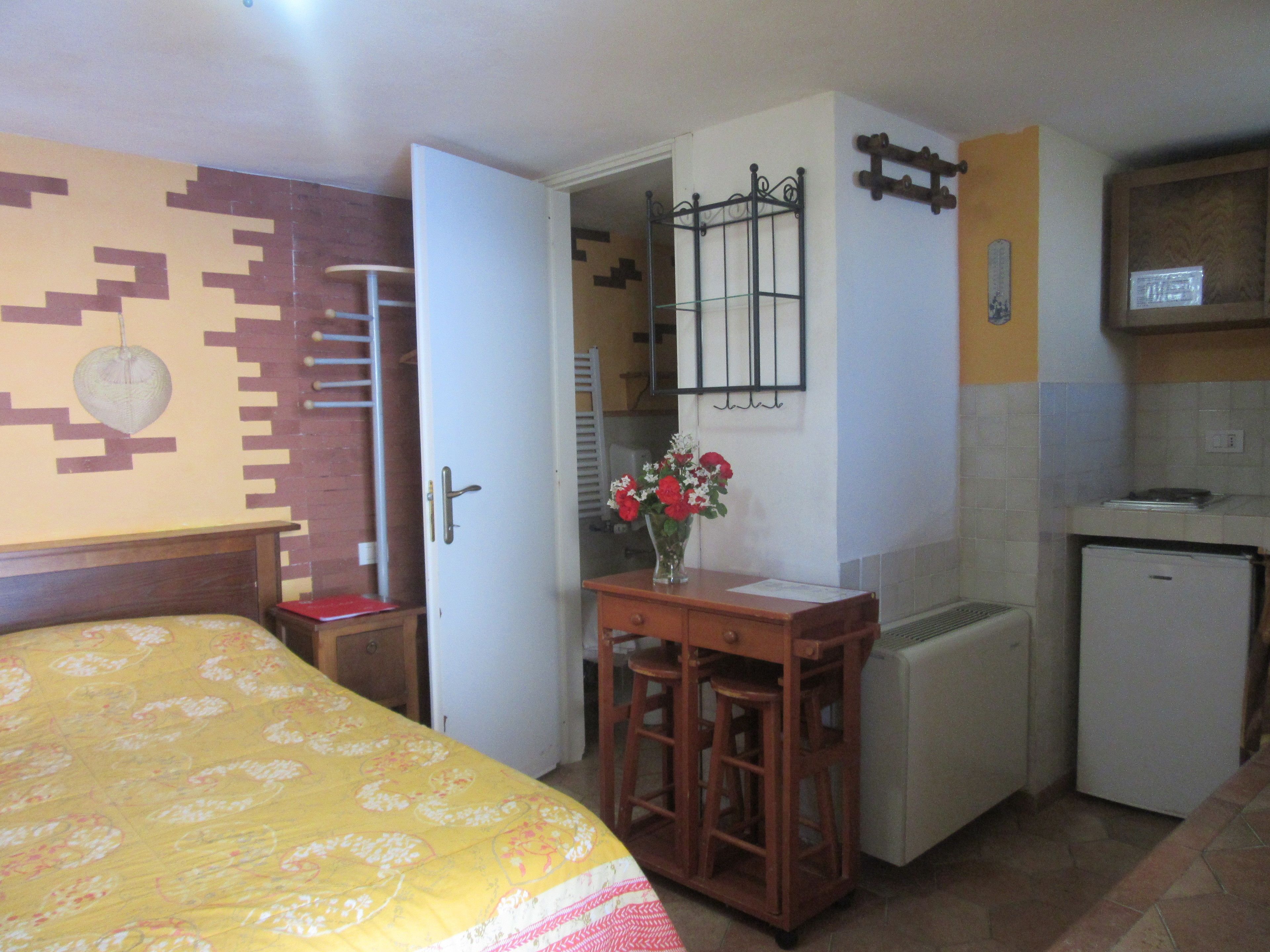 Una habitación compacta con cama, una mesita con taburetes, refrigerador y espejo en la pared.