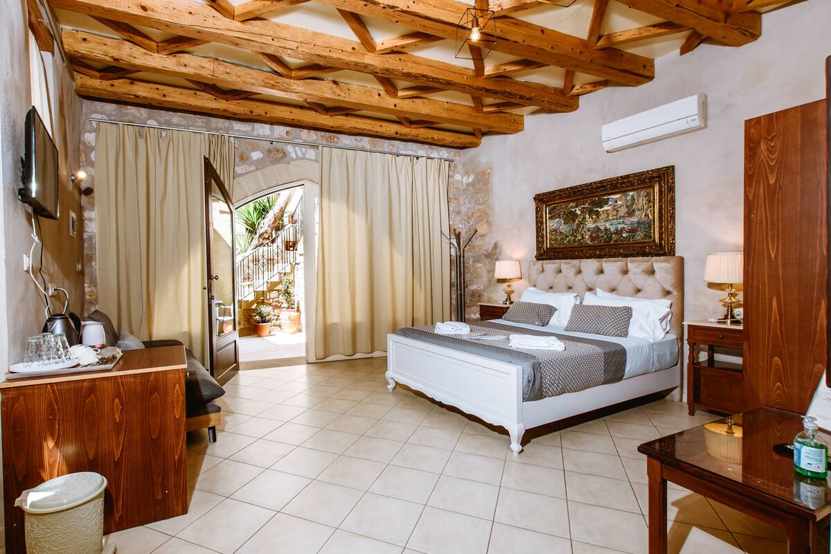 A bedroom with a bed, wooden ceiling beams, a painting, and a view through a door.