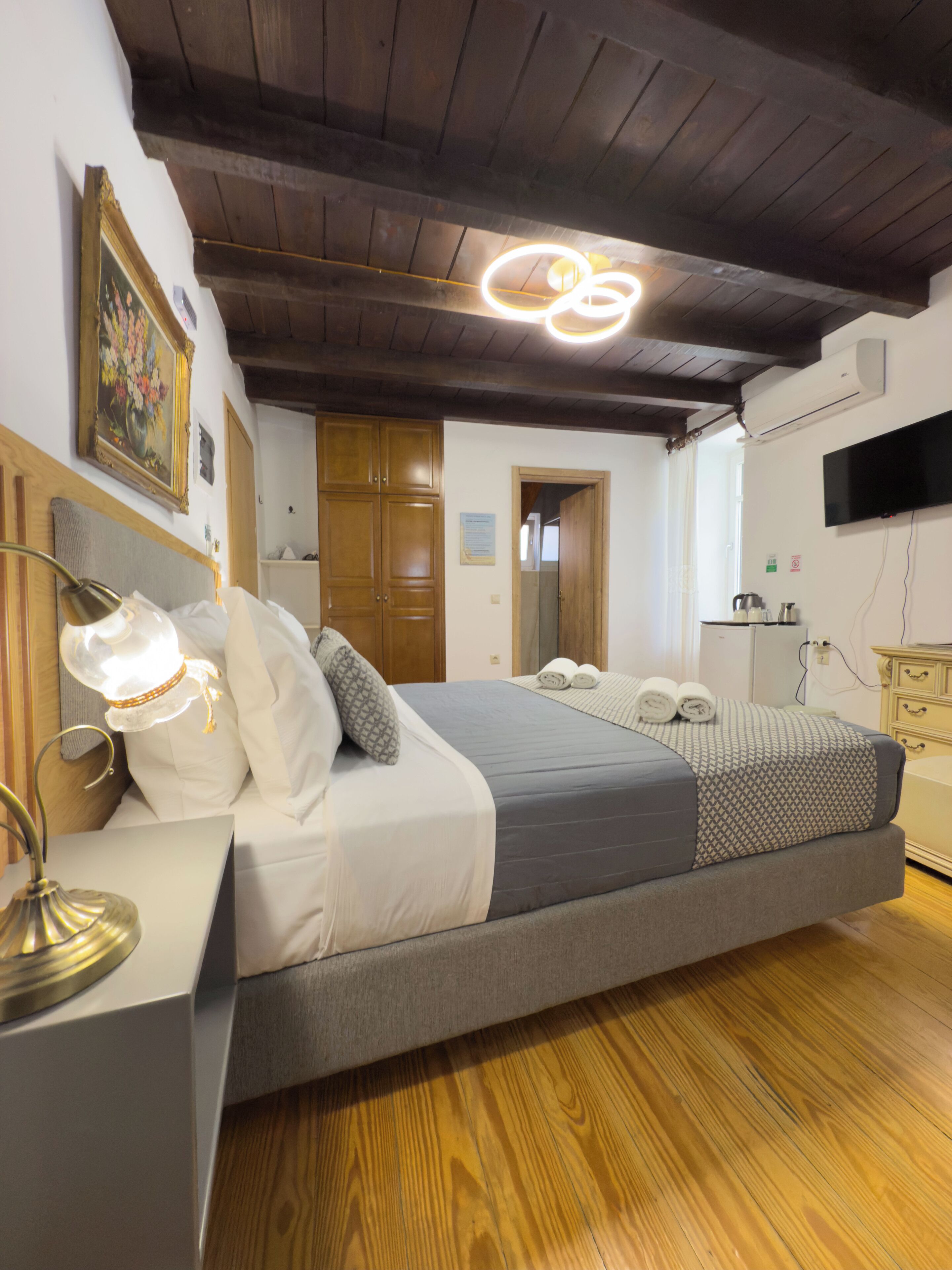 Quarto com teto de madeira, cama com roupa de cama cinza, abajur, pintura na parede e televisão na parede.