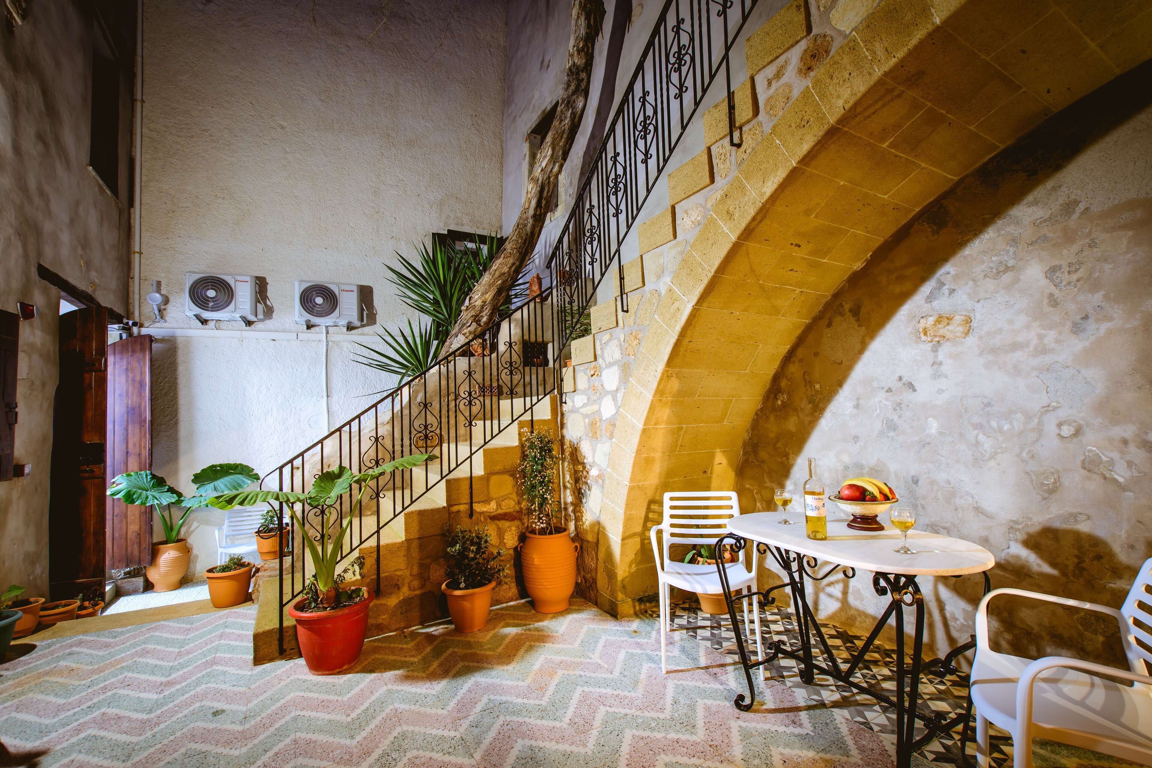 A courtyard with a staircase, potted plants, a table with chairs, and a rustic stone wall.