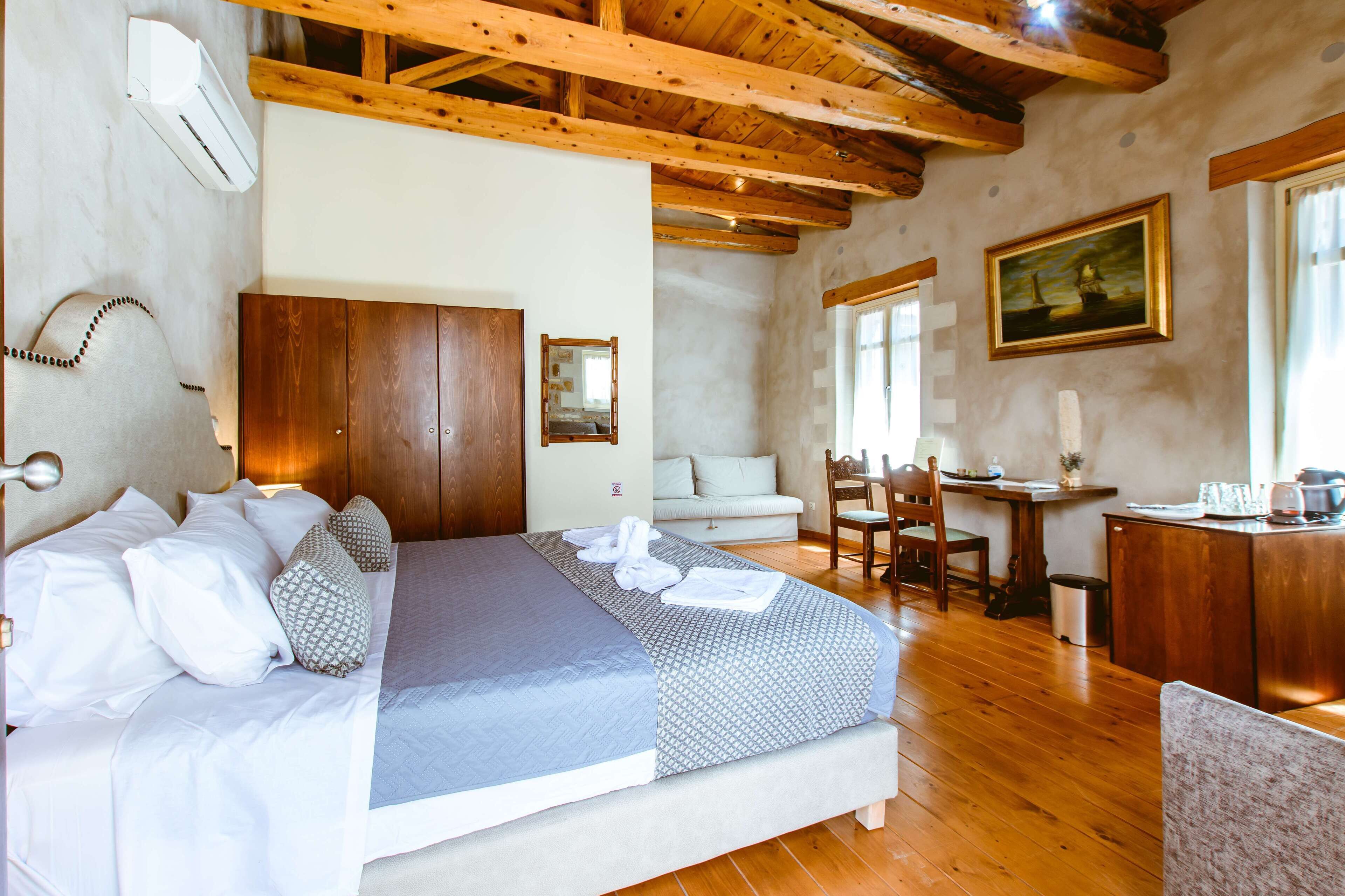 Un dormitorio con una cama grande, vigas de madera en el techo, un armario de madera, un escritorio con una silla y un cuadro en la pared.