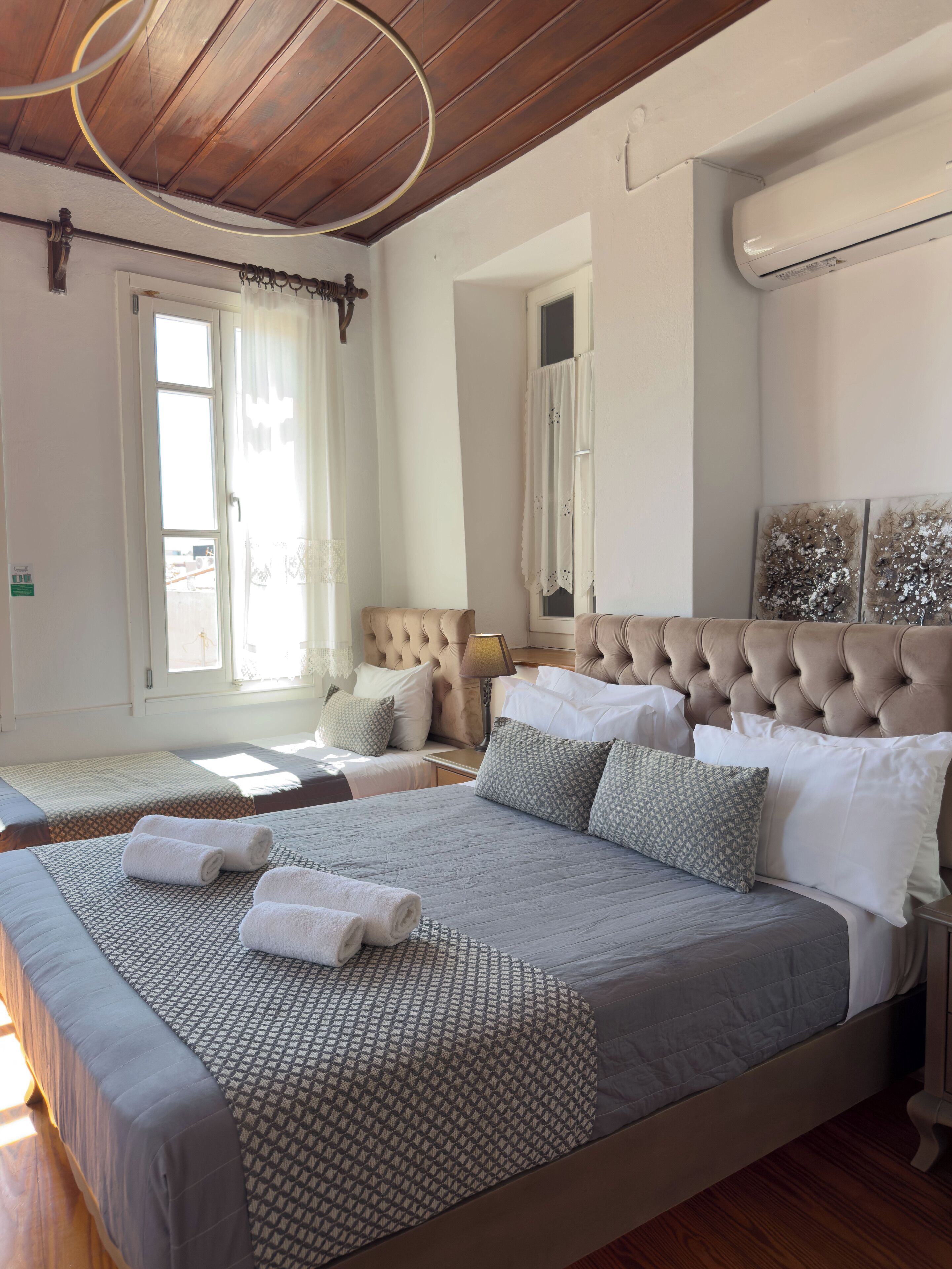 A hotel room with a large bed, a smaller bed, and a sofa. There is a window with curtains, a ceiling fan, and a wall-mounted air conditioner.