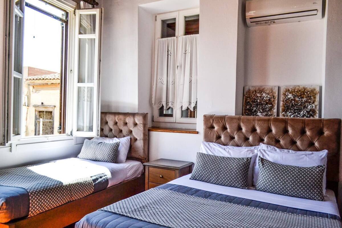 A hotel room with two beds, a window with curtains, an air conditioning unit, and a view of the outside.
