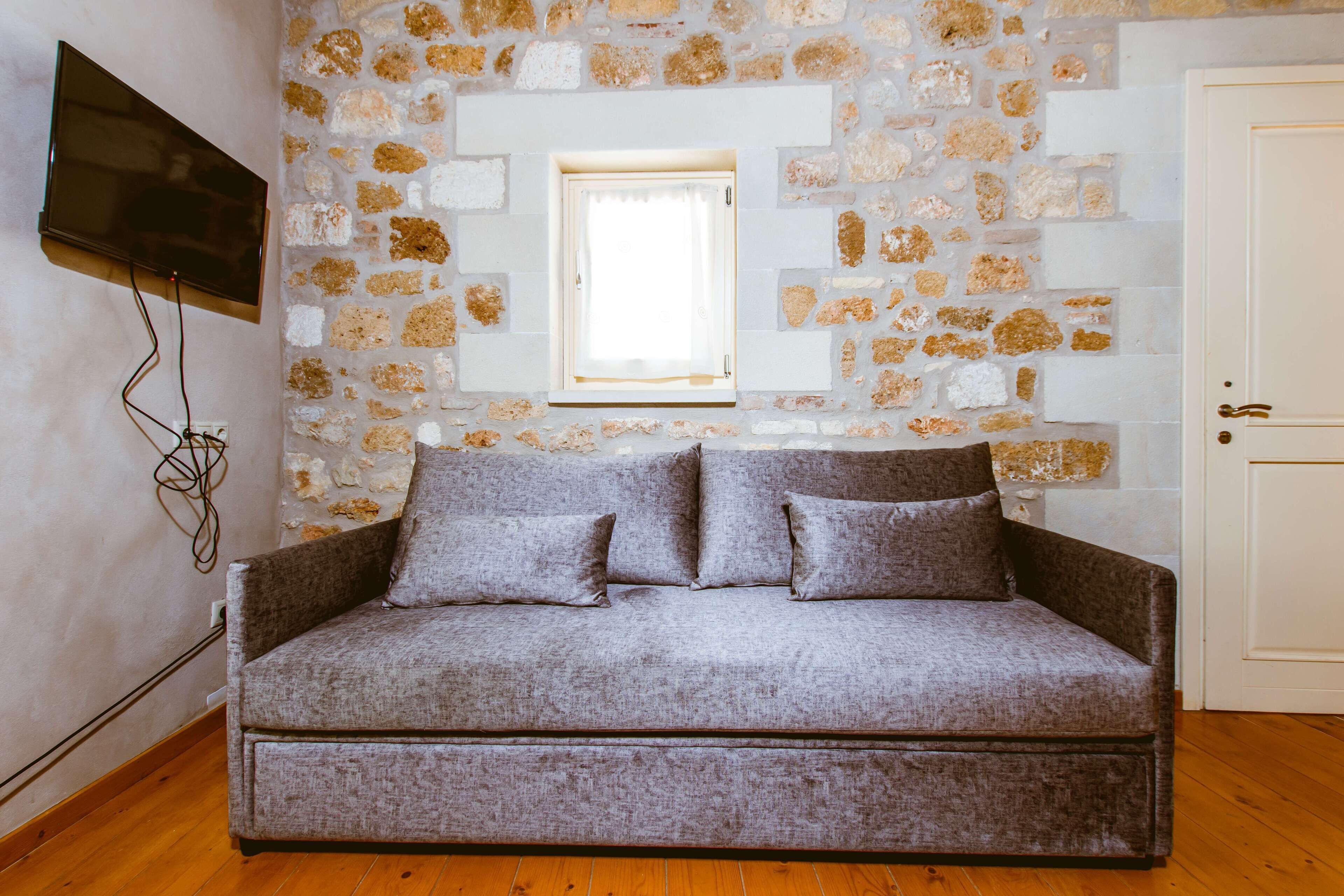 Une pièce avec un mur en pierre, une fenêtre, un canapé gris et un téléviseur fixé au mur.