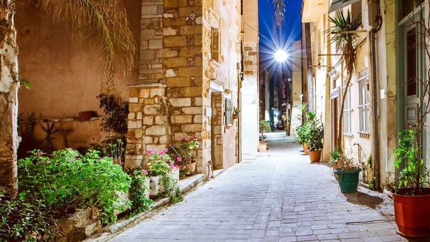 Uma rua de paralelepípedos estreita, ladeada por edifícios com fachadas de pedra e plantas em vasos.