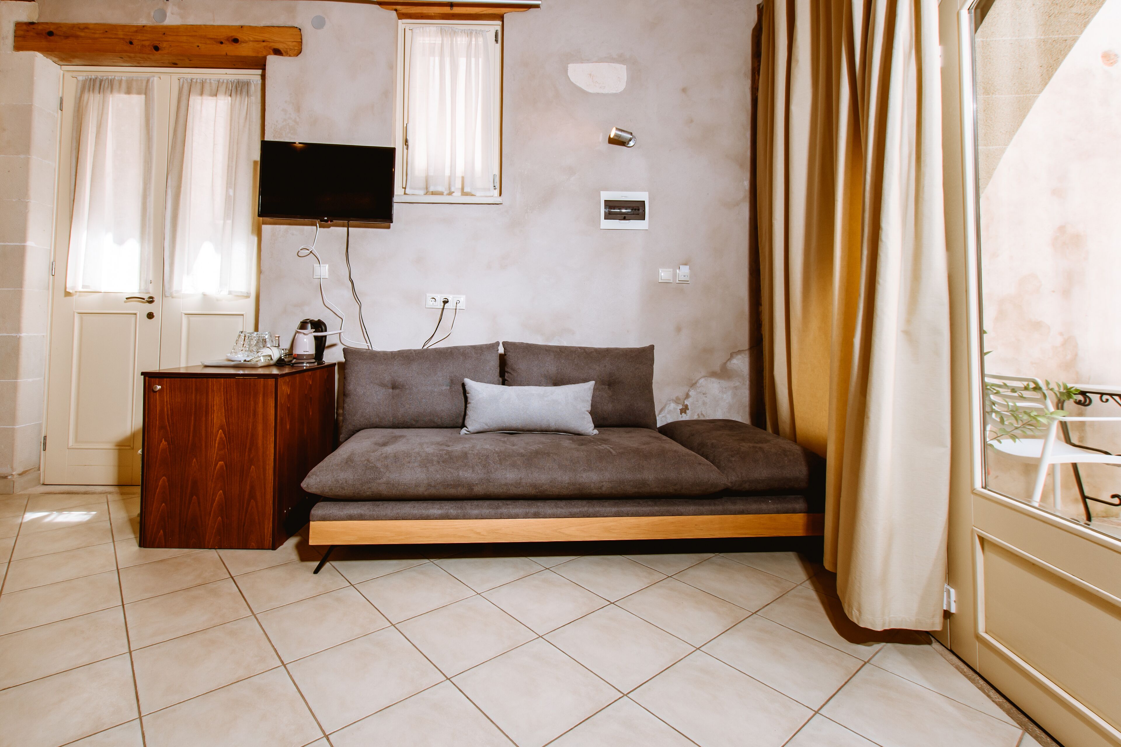 A room with a sofa, a wooden side table, a television mounted on the wall, and a window with curtains.