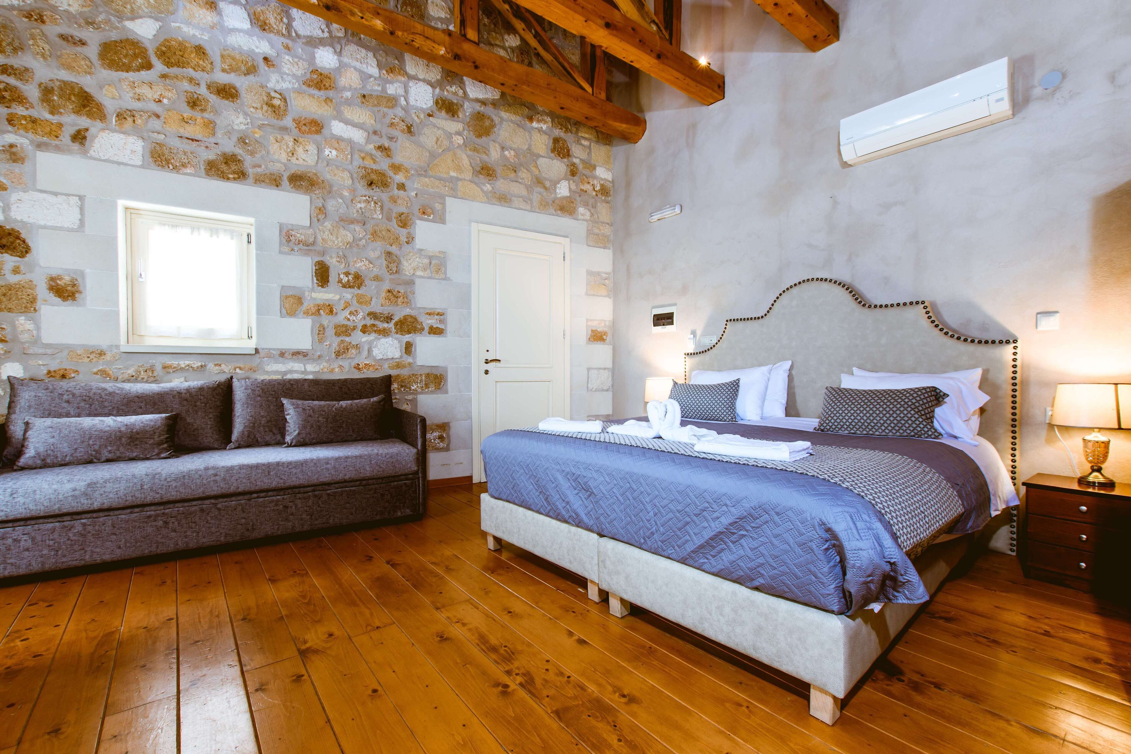 A bedroom with a stone wall, wooden floor, a bed with a headboard, a sofa, and a nightstand with a lamp.