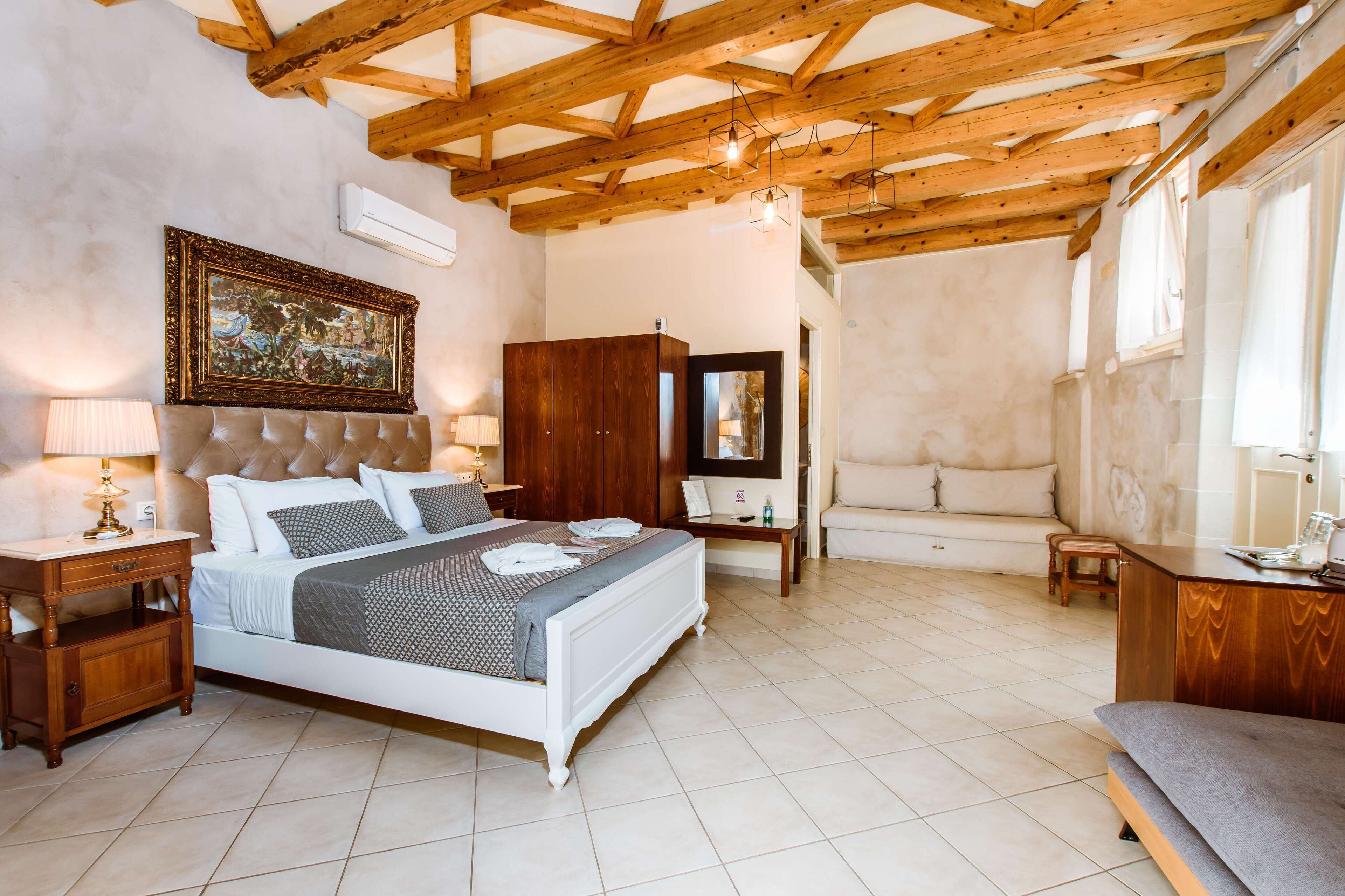 A bedroom with a large bed, wooden ceiling beams, a painting, a sofa, and a desk.