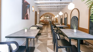 A modern restaurant interior with high ceilings, geometric patterned flooring, and a central archway leading to the kitchen. Tables are arranged in rows with chairs, and pendant lights hang from the ceiling. - El Greco (Chania)