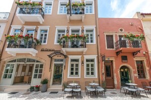 Ένα κτίριο με πολλά μπαλκόνια στολισμένα με λουλο ύδια, έναν χώρο καθιστικού στο ισόγειο με τραπέζια και καρέκλες και μια ευδιάκριτη είσοδο με μια επιγραφή.