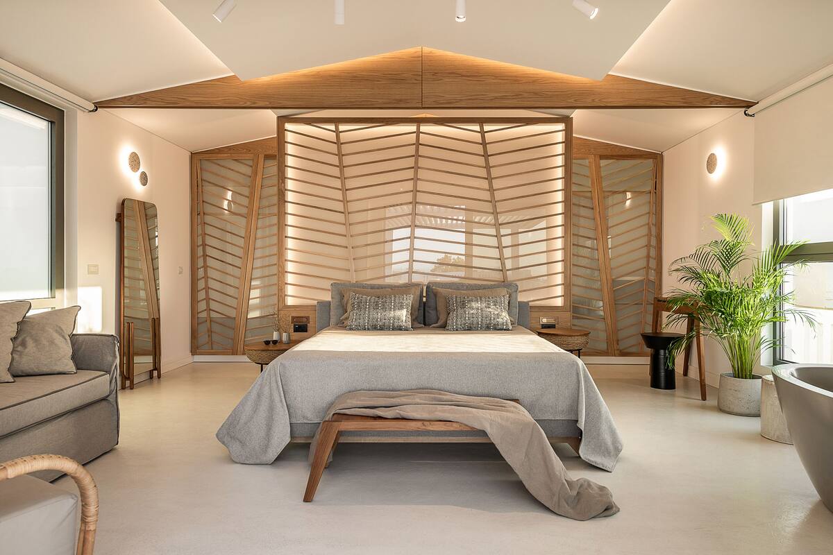 A modern bedroom with a large bed, wooden slats, a grey sofa, and a potted plant.