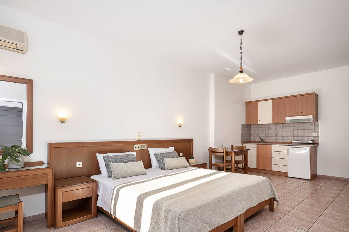 A hotel room with a bed, bedside tables, a desk, a chair, a small table, a hanging light fixture, and a kitchenette.