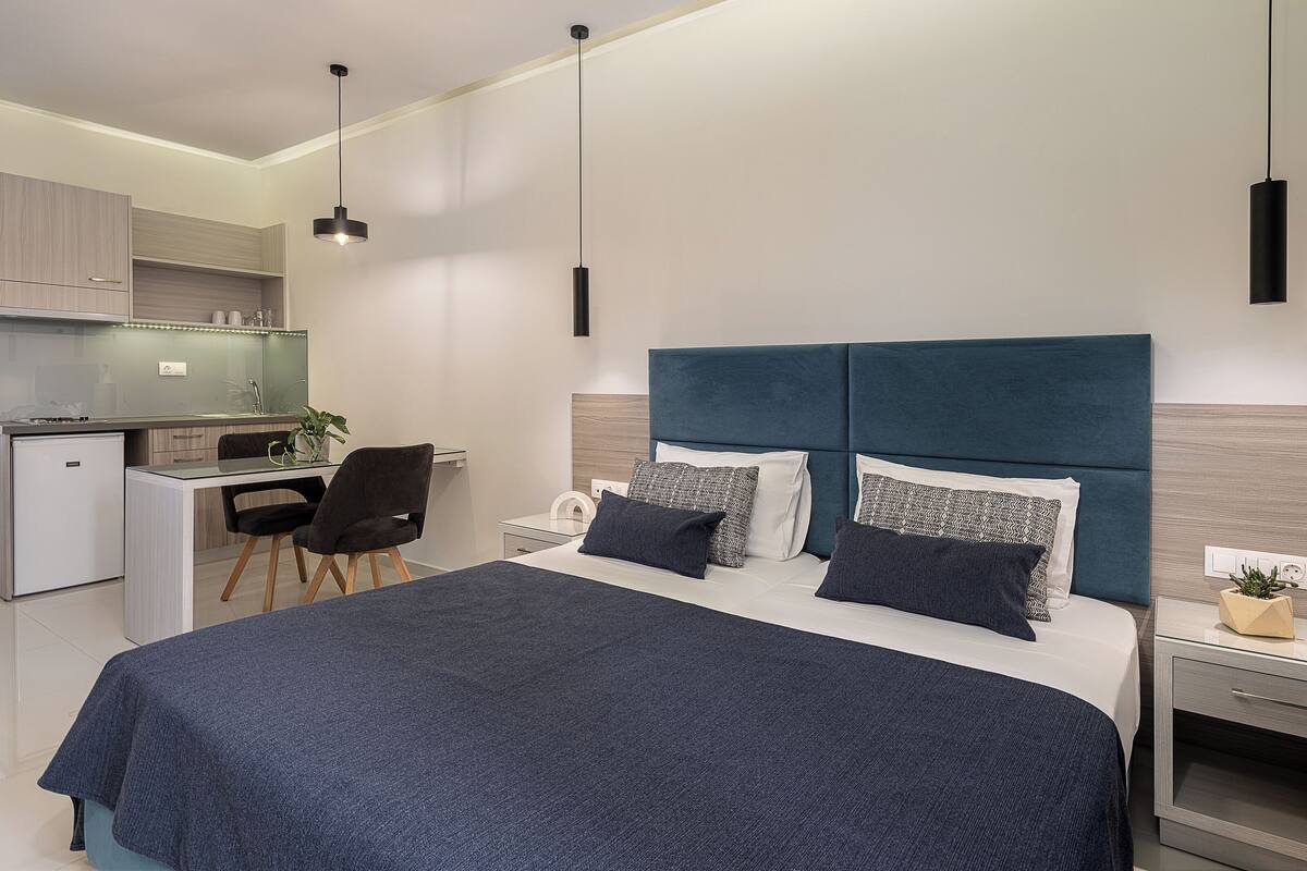 A modern bedroom with a large bed, bedside tables, a desk, and a kitchenette.