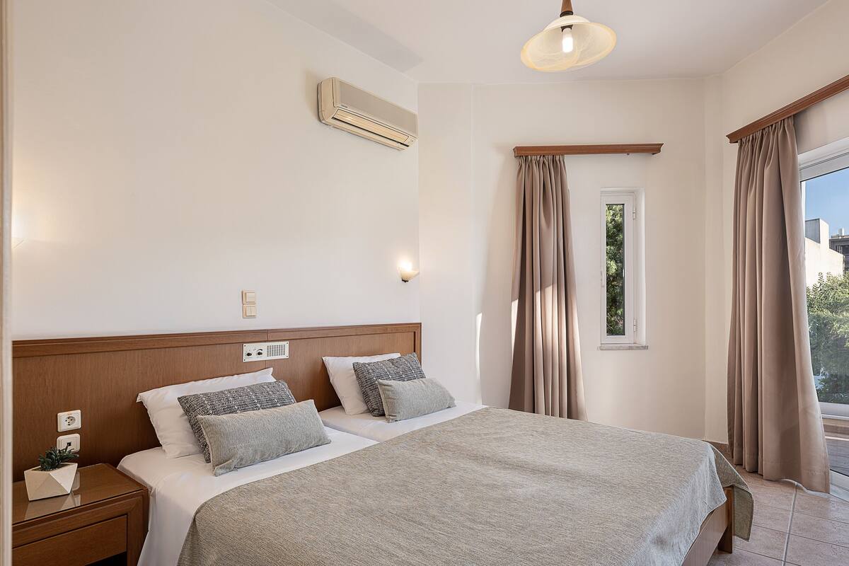 A hotel room with a bed, nightstand, bedside table, lamp, air conditioning unit, and a window with curtains.