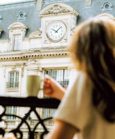 Una persona sosteniendo una taza, con vista a un edificio con un reloj y arquitectura ornamentada.