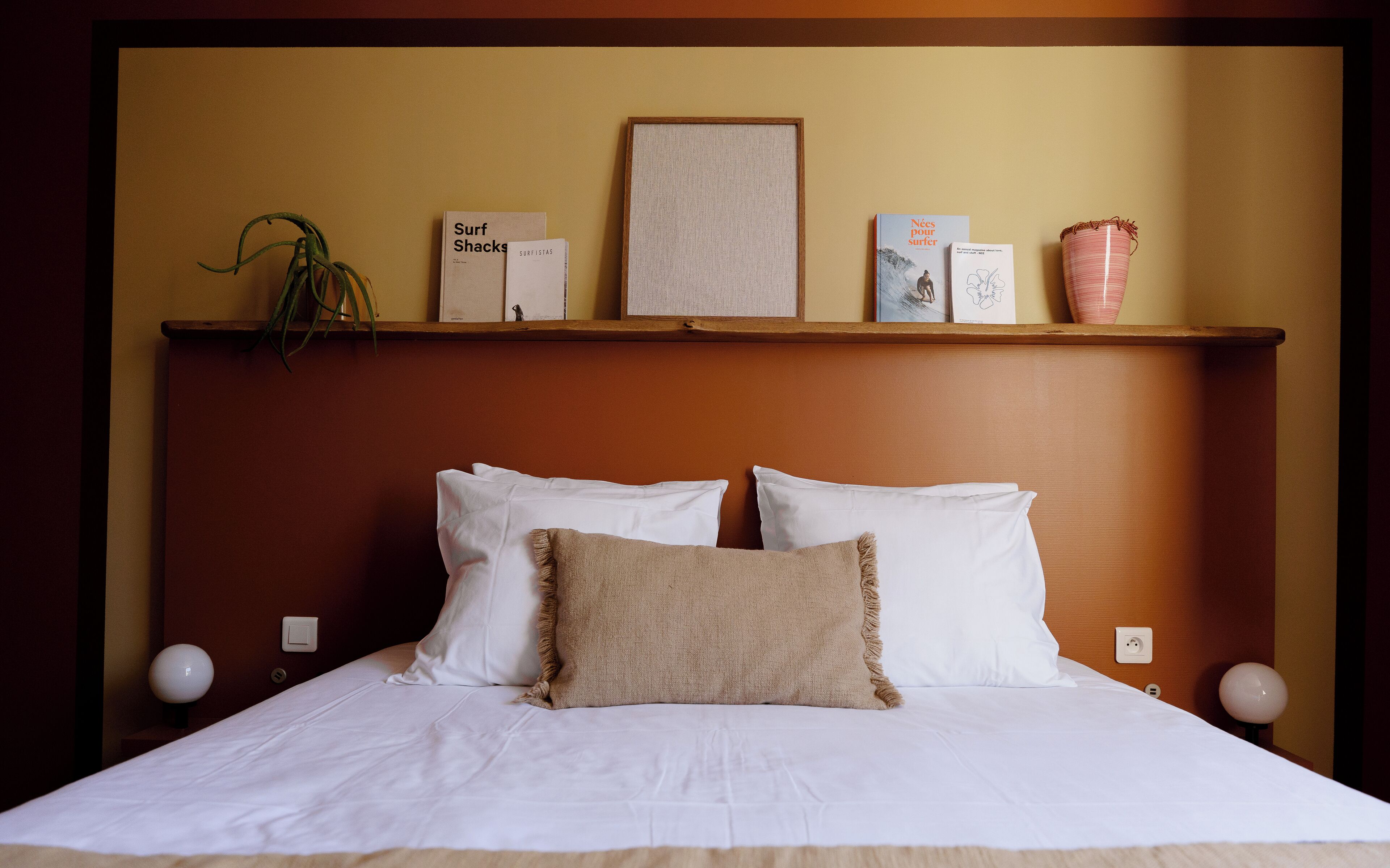 A hotel room with a bed, white pillows, a beige throw, a shelf with books, a framed picture, and a potted plant.