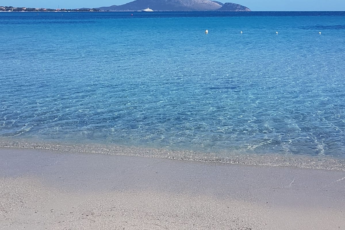 En strand med kristallklart blått vatten, en sandstrand och ett berg i fjärran.