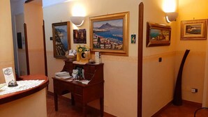 A hotel lobby with a wooden reception desk, a small seating area, and framed coastal landscape paintings on the walls. - Conte Cavour (Naples)