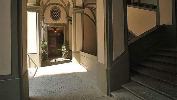 A hallway with a staircase leading to a door with a window. There are potted plants and a light fixture on the wall. - Conte Cavour (Naples)