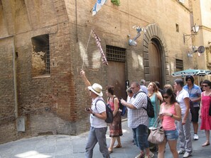A group of people walking on a cobblestone street in front of a historic building with brick walls and an arched doorway. - Palazzo Masi (Siena)