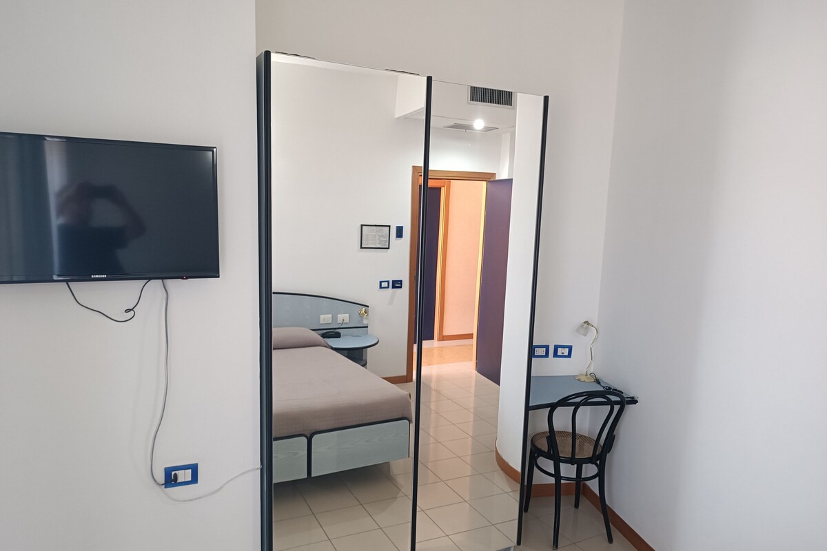 A hotel room with a bed, a desk, a chair, a TV mounted on the wall, and a mirror.