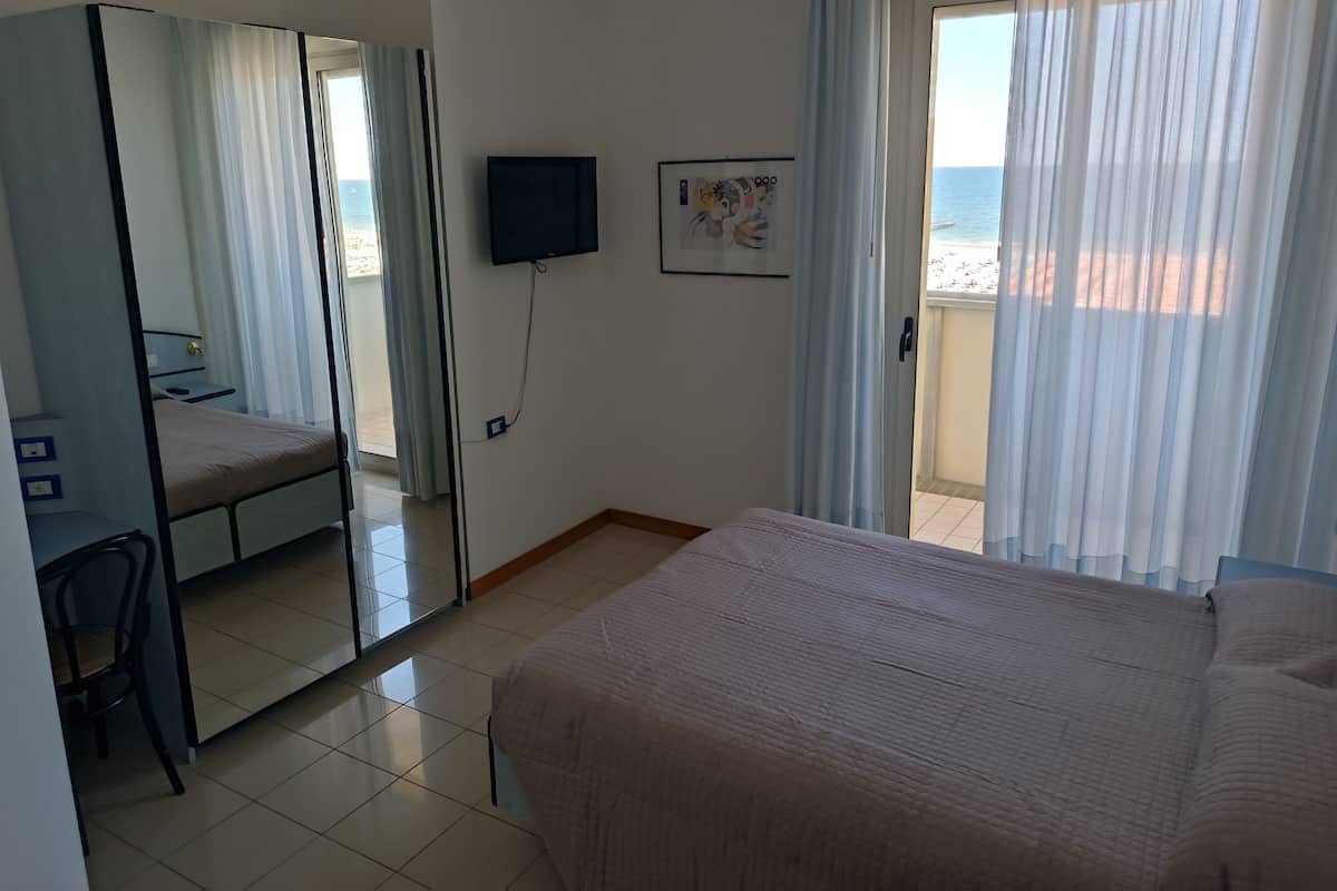 A hotel room with a bed, a desk, a chair, a TV, and a balcony with a view of the beach.