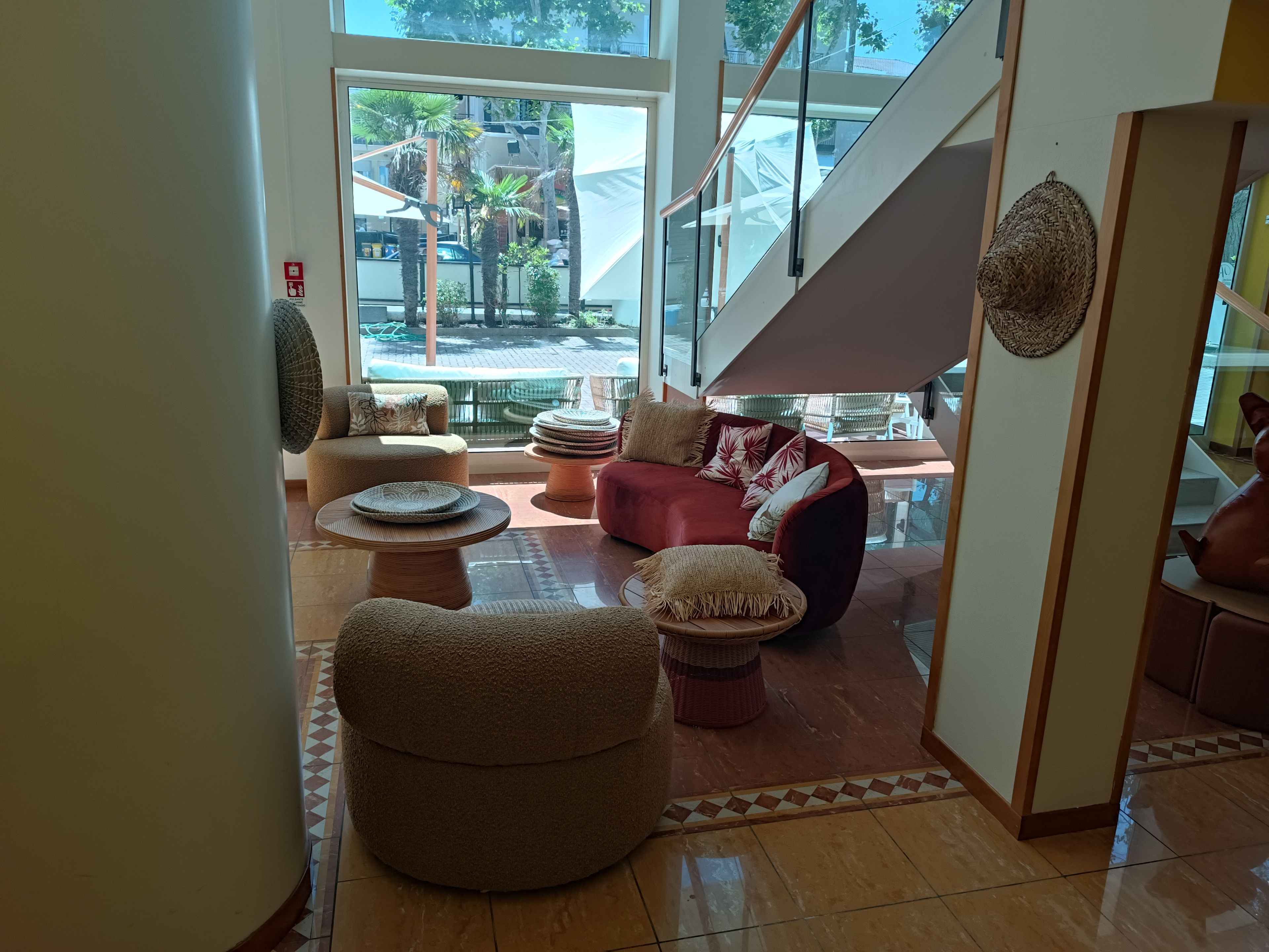 Un salón amplio con un sofá rojo, varios otomanes redondos y un ventanal con vistas a la calle y palmeras.