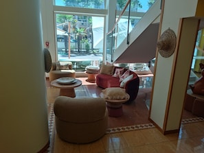 A spacious living area with a red sofa, multiple round ottomans, and a large window offering a view of the street and palm trees. - Remin Plaza (Rimini)