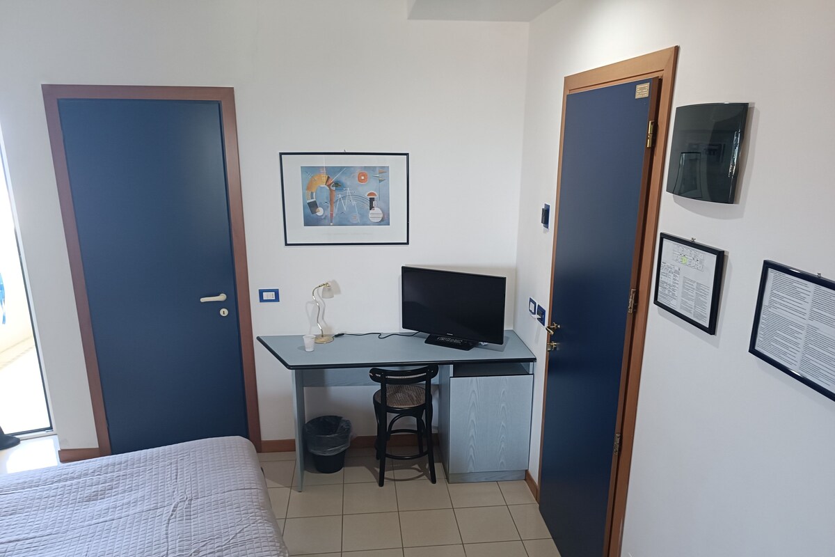 A small, compact room with a bed, a desk with a computer, a chair, and a trash bin. There are two doors, one on each side of the room.