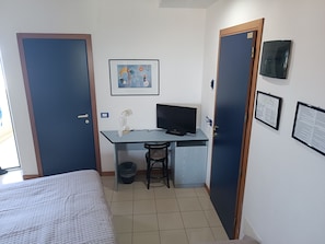 Ein kleiner, kompakter Raum mit einem Bett, einem Schreibtisch mit Computer, einem Stuhl und einem Mülleimer. Es gibt zwei Türen, eine an jeder Seite des Raumes.