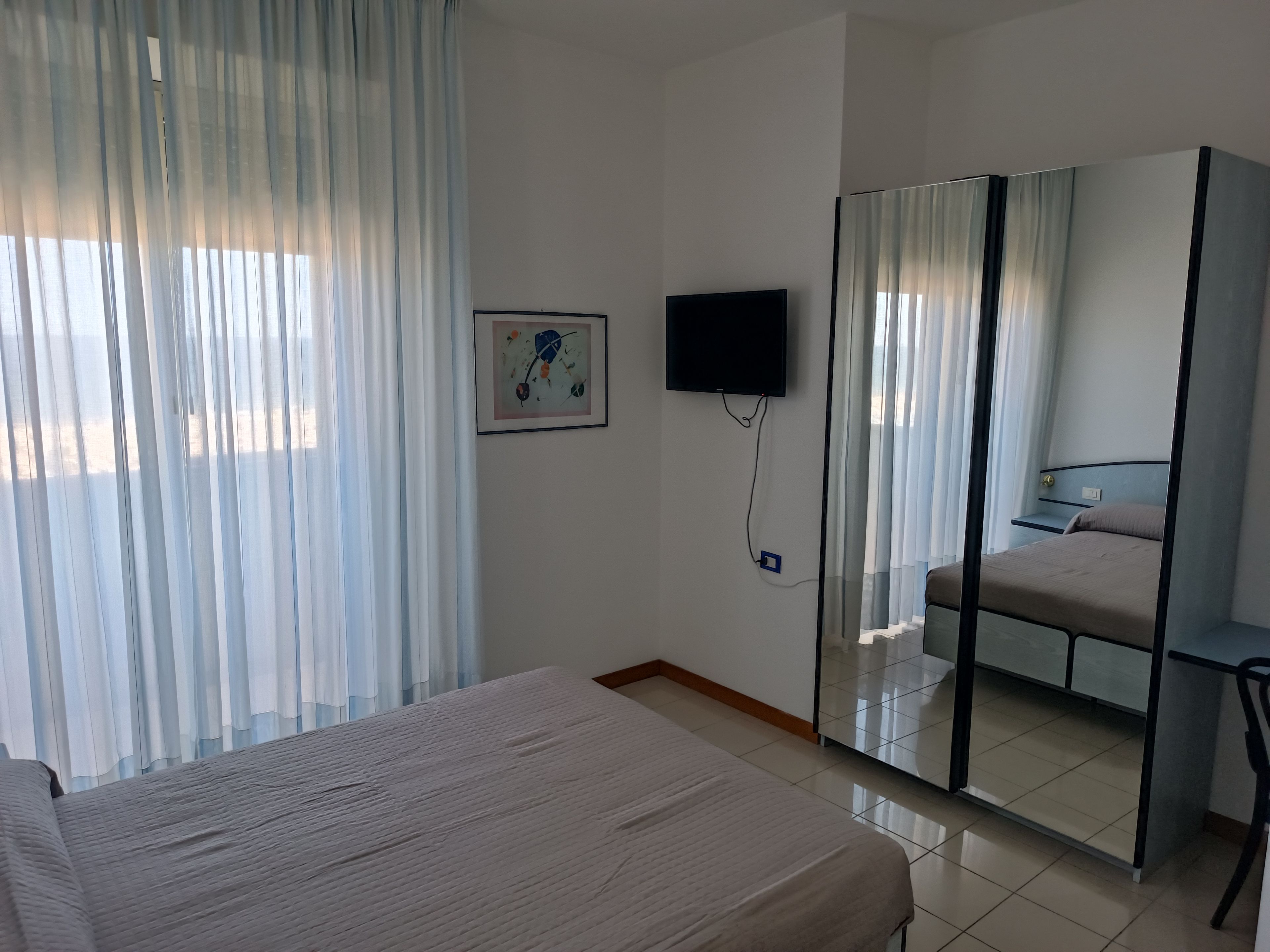 Habitación de hotel con cama, televisor montado en la pared, espejo y ventana con cortinas translúcidas.