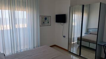Habitación de hotel con cama, televisor montado en la pared, espejo y ventana con cortinas translúcidas.