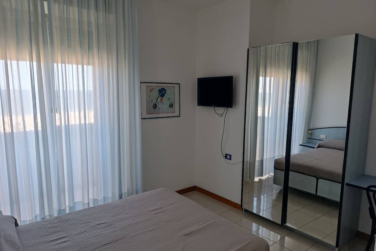 A hotel room with a bed, a TV mounted on the wall, a mirror, and a window with sheer curtains.