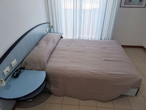 A neatly made bed with a beige cover, a small bedside table with a lamp, and a power outlet on the wall. - Remin Plaza (Rimini)