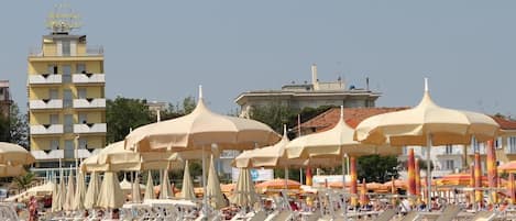 Una playa con filas de tumbonas y sombrillas, un edificio de hotel al fondo y cielos despejados.