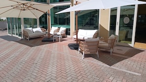 En udendørs terrasse med fletmøbler og hvide hynder, en glasdør indgang og et pool, der kan ses gennem glasset.