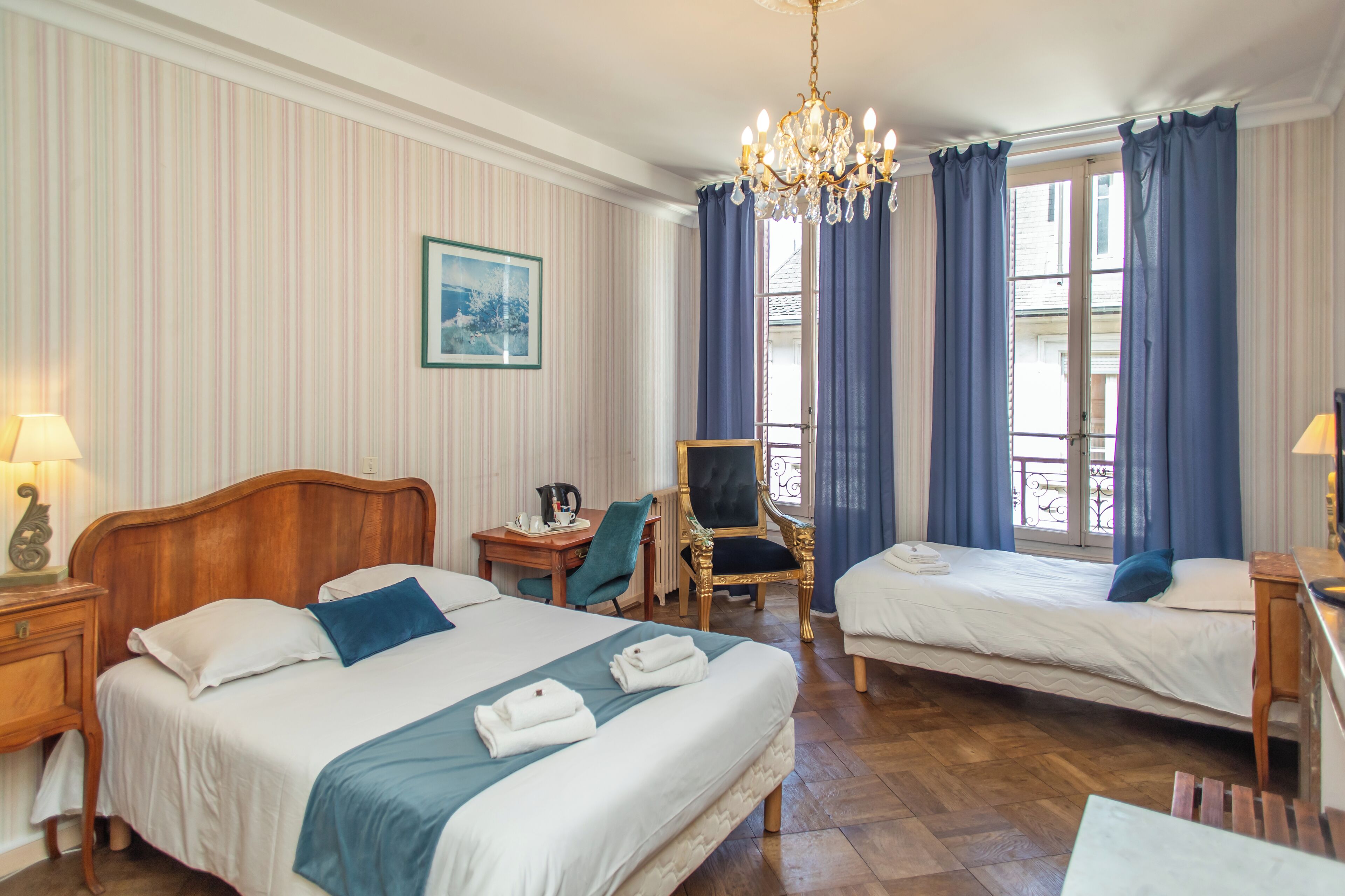 A hotel room with two beds, a desk, a chair, a chandelier, and a window with curtains.