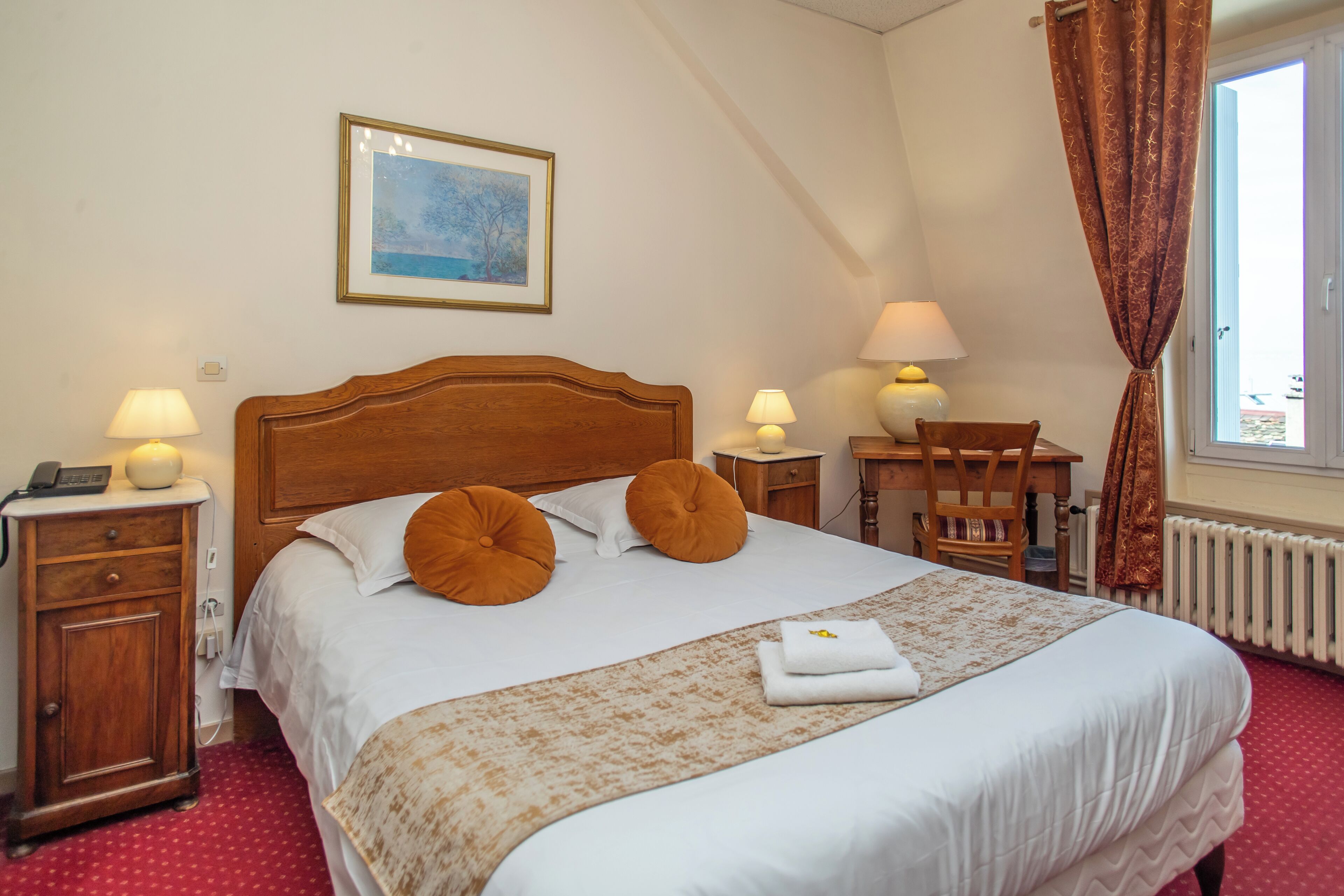 Een opgemaakt bed met kussens, een nachtkastje met lamp, een stoel en een raam met gordijnen.