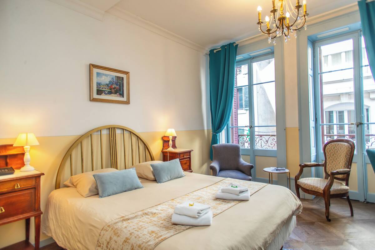 Hotellihuone, jossa on suuri sänky, kaksi tuolia, pieni pöytä, lamppu ja taulu seinällä.