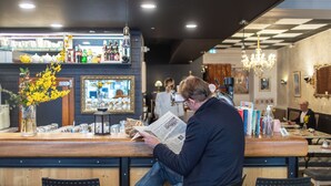 Un homme est assis au bar, lisant un journal. Le bar est doté d’un comptoir en bois, d’étagères remplies de bouteilles et d’un lustre.