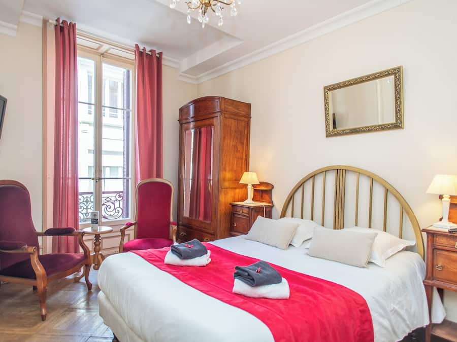 A hotel room with a bed, two chairs, a wooden wardrobe, a desk, a lamp, and a television.